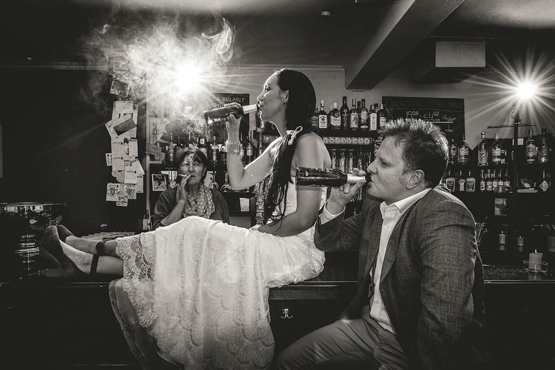 Braut liegt im Brautkleid lachend auf einer Bar, der Bräutigam steht daneben