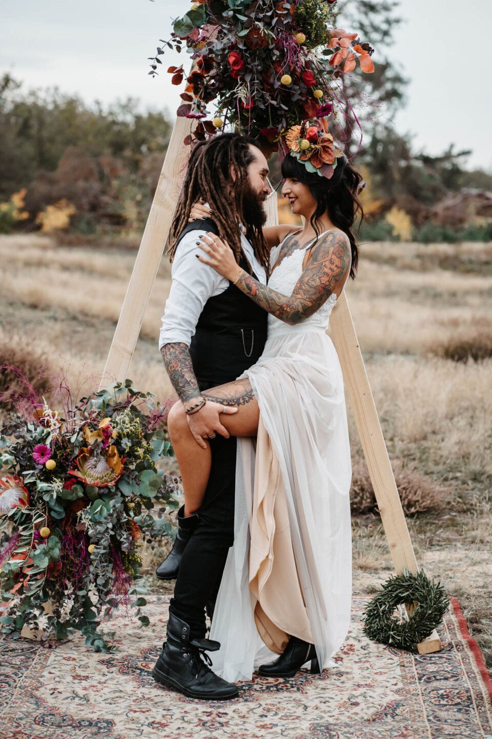 Braut mit Tattoos umramt Bräutigam vorm Traubogen