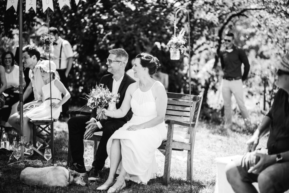Brautpaar auf einer Bank auf der Wiese vorm Trauversprechen bei ihrer Gartenhochzeit