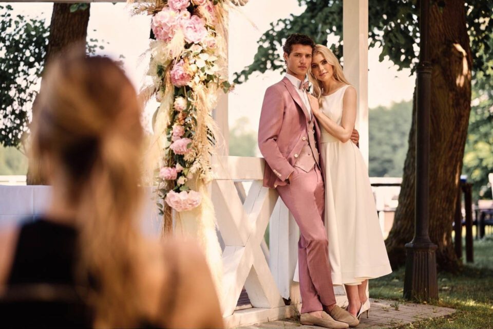 Braut mit köchellangem Brautkleid lehnt sich an Bräutigam im Roséfarbenen Anzug