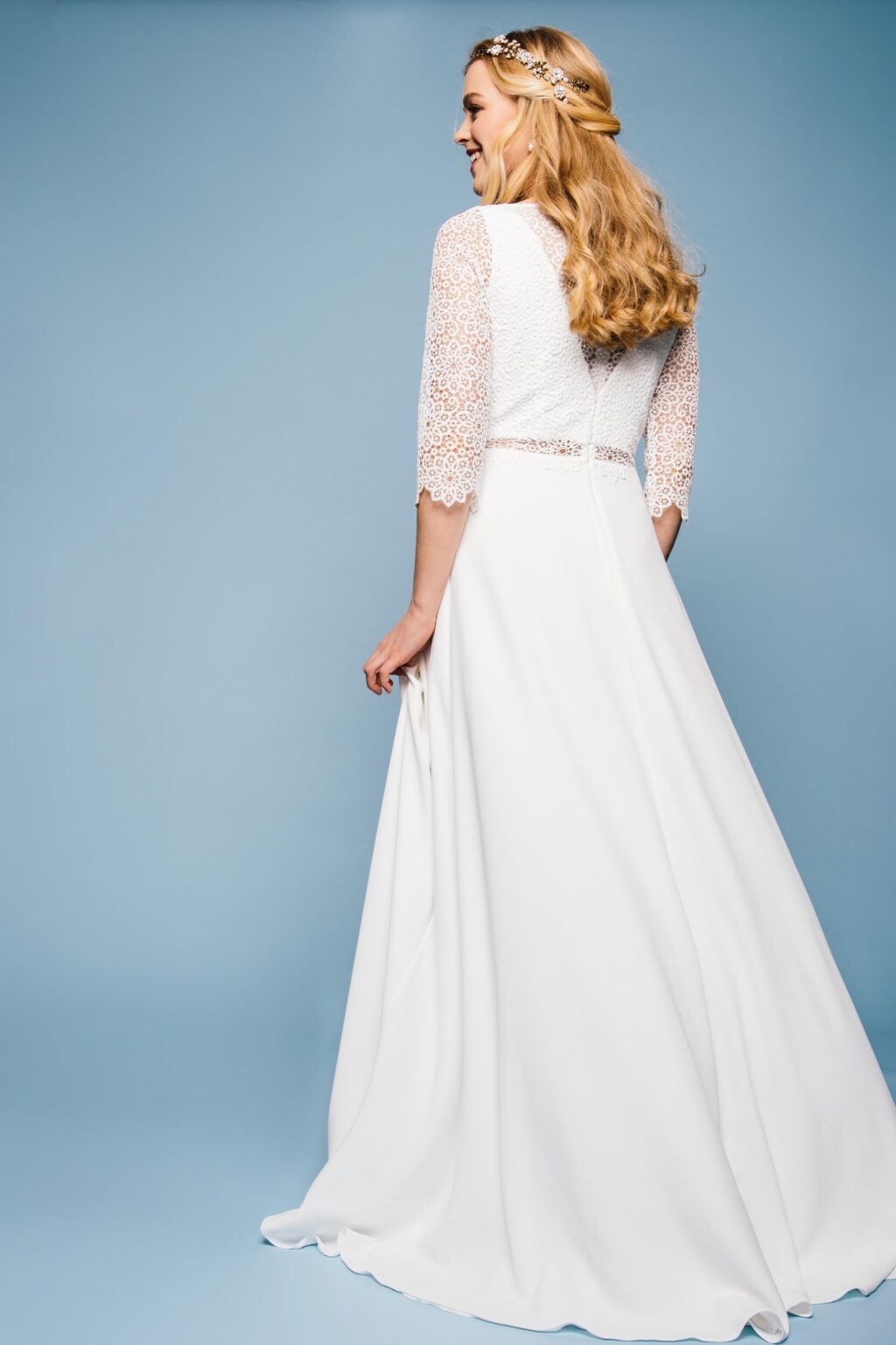 Braut von hinten im Spitzenkleid mit langen Ärmeln vor blauem Hintergrund