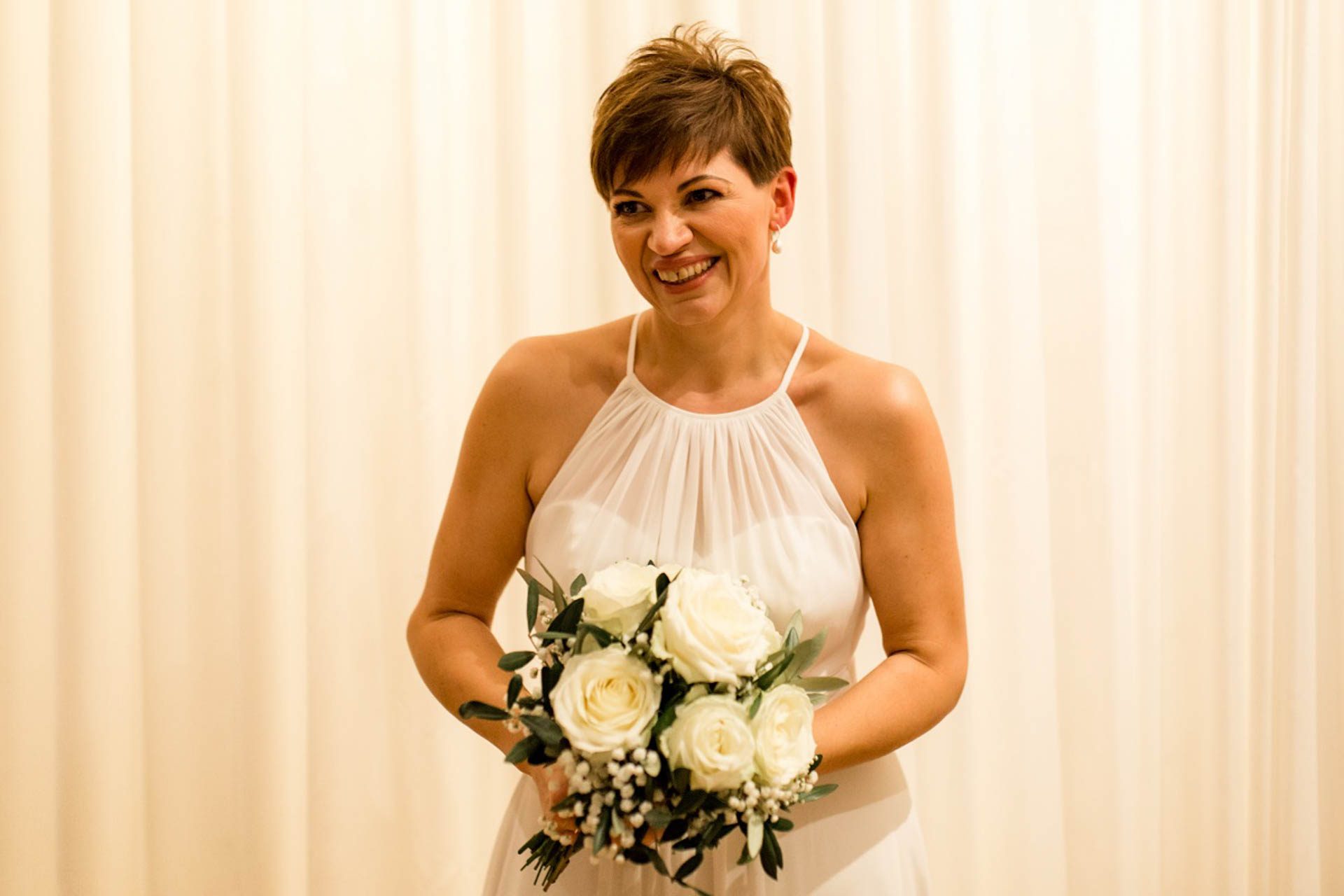 Lachende Braut mit kurzen Haaren im schlichten Brautkleid KUESSdieFINJA mit weißem Brautstrauß