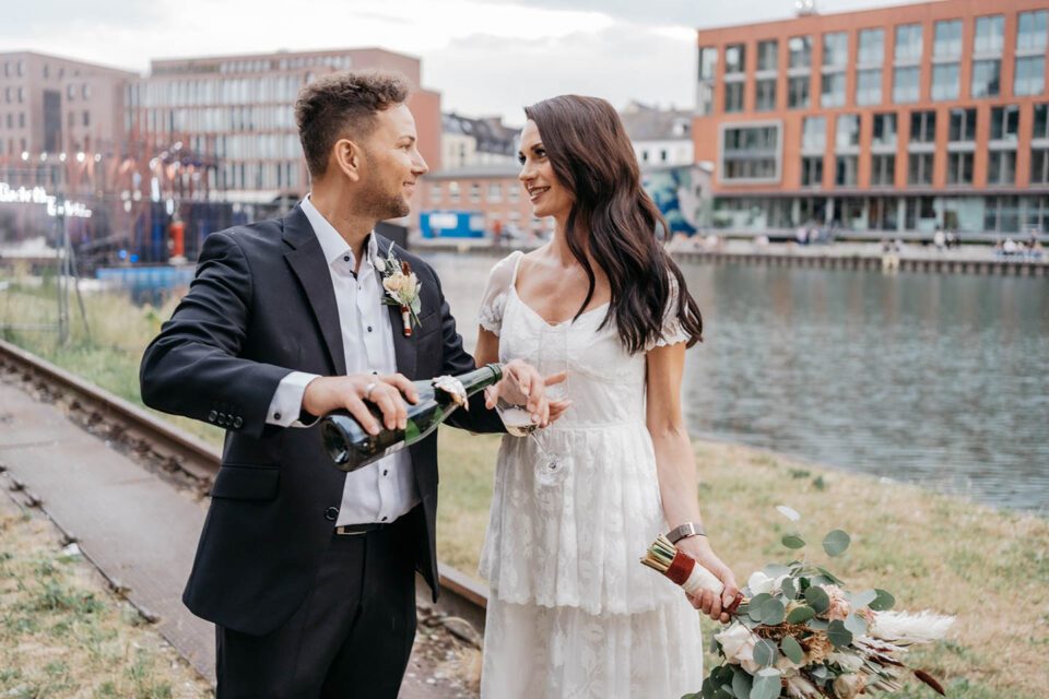Bräutigam gießt in Münsters Hafen Sekt ein und lächelt Braut im Brautkleid mit Stufenrock dabei an