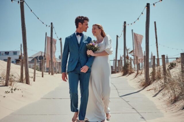 Braut im Puffärmel Brautkleid hakt sich bei Bräutigam ein und schlendert barfuß am Strandweg