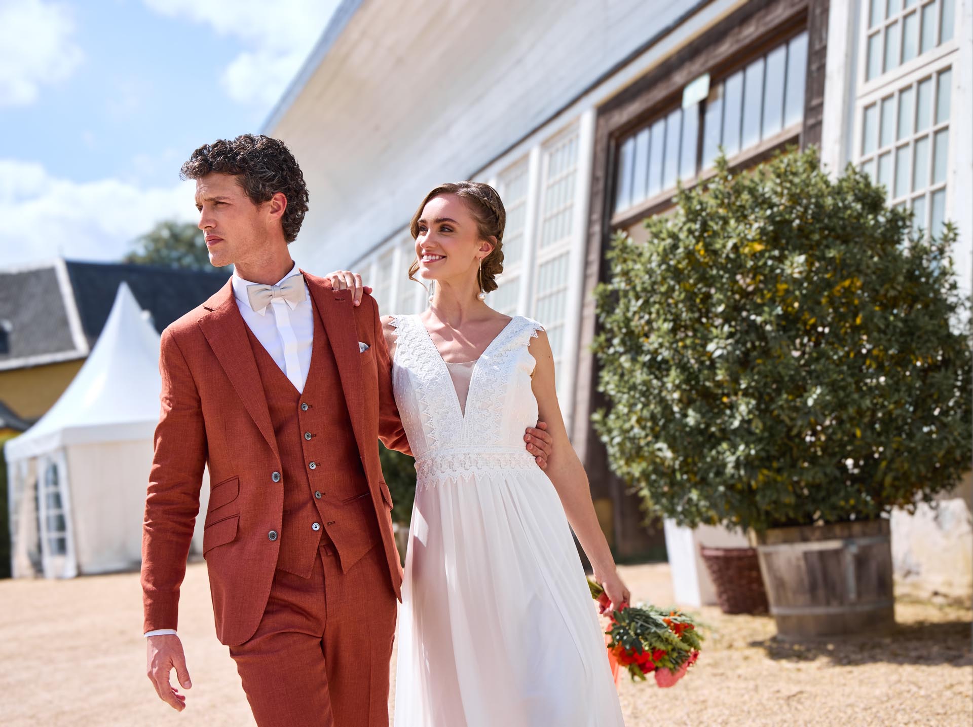 Modernes Brautkleid läuft Arm in Arm im Freien. Braut trägt ein taillenbetontes Brautkleid im nachhaltigen Materialmix, der Bräutigam einen rotbraun melierten Hochzeuitsanzug.