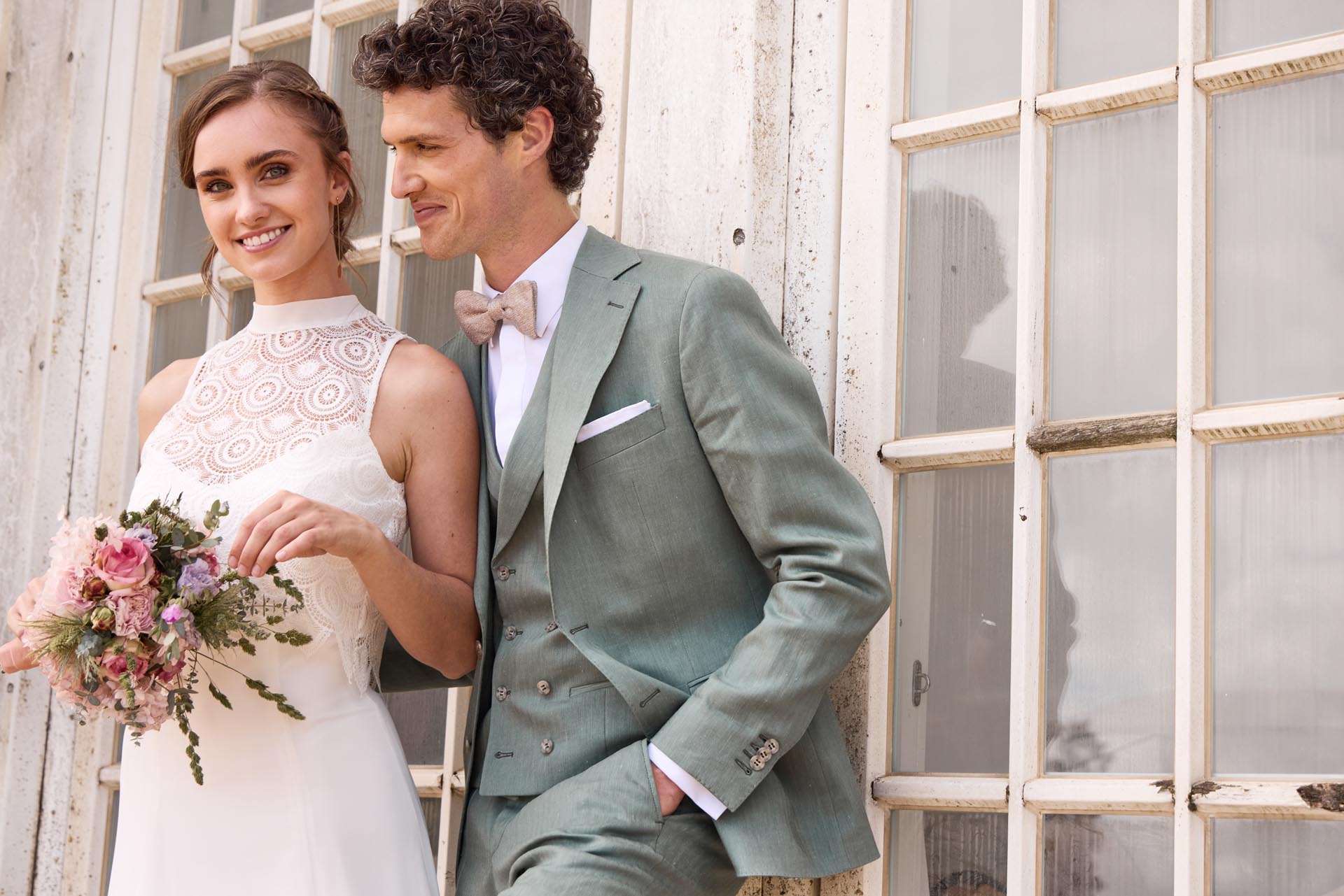 Wunderschön lächelnde Braut im Stehkragen Brautkleid mit Brautstrauß neben Bräutigam mit rosa Fliege und hellolivem Anzug.