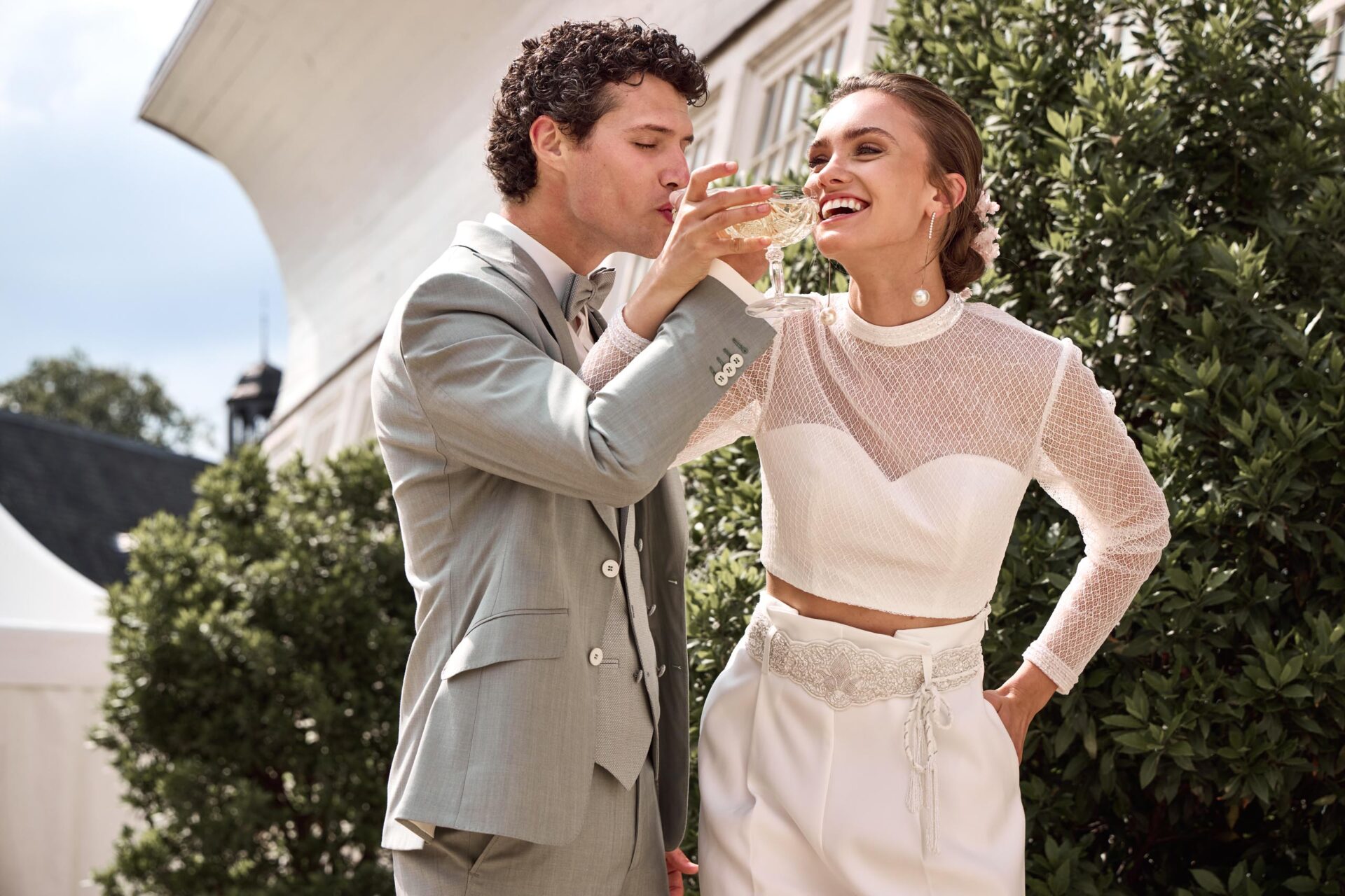 Brautpaar trinkt Champagner aus Schalen und hat die Arme mit den Gläsern dabei überkreuzt. Braut in Hochzeitshose und schimmerndem Langarmoberteil, Bräutigam in hellen Grünoliven Hochzeitsanzug