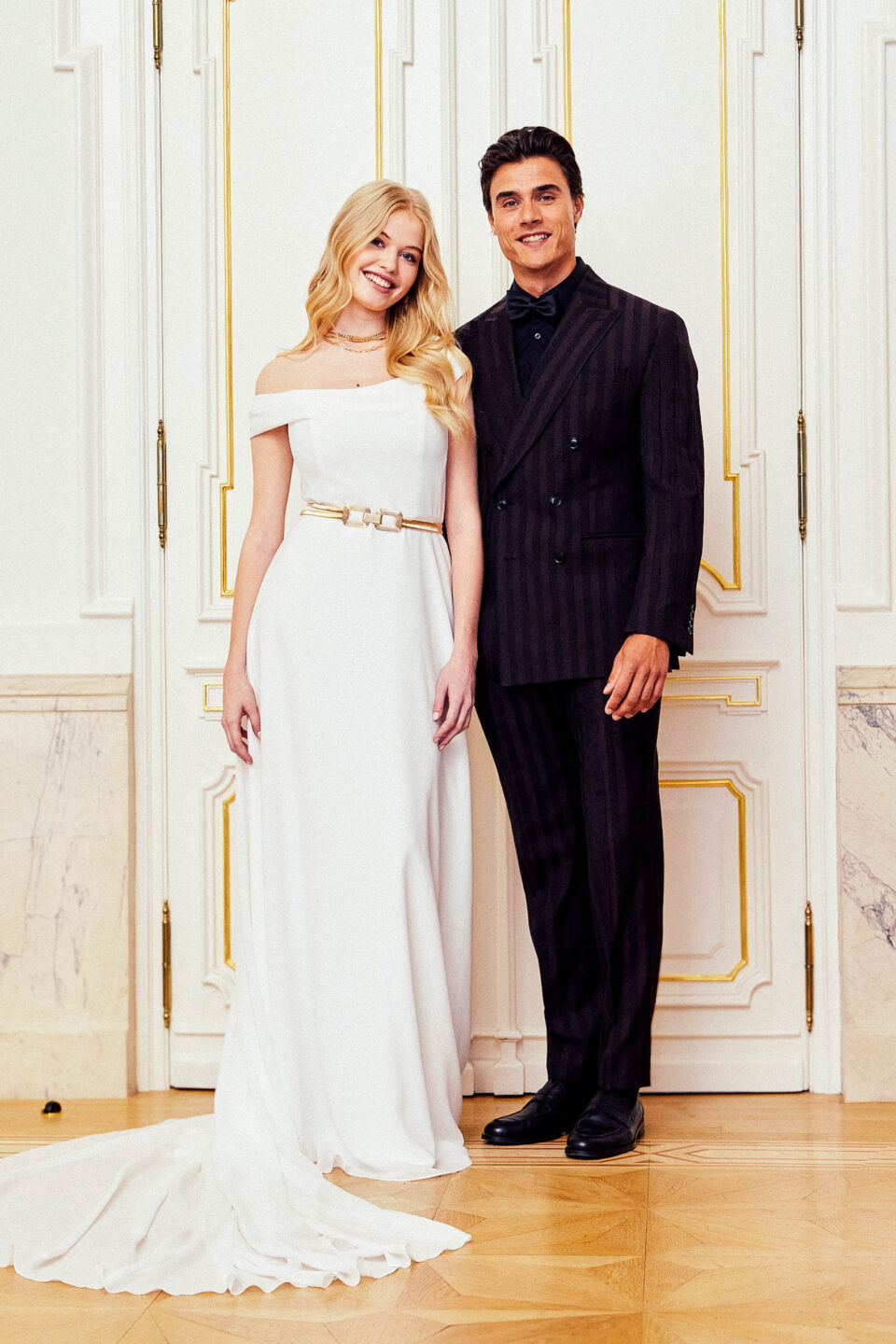 Schmales Chiffon-Brautkleid KUESSdieCARLOW mit asymmetrischem Carmenausschnitt und goldenem Gürtel, Brautpaar im Palais