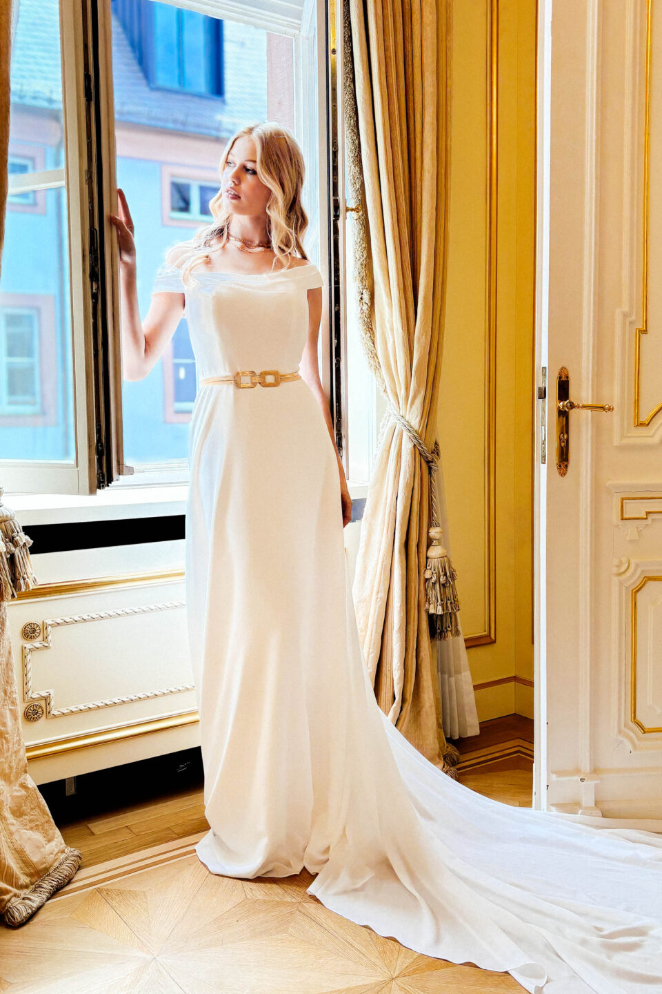 Brautkleid Carlow mit schulterfreiem Carmenausschnitt und Schleppe am Fenster