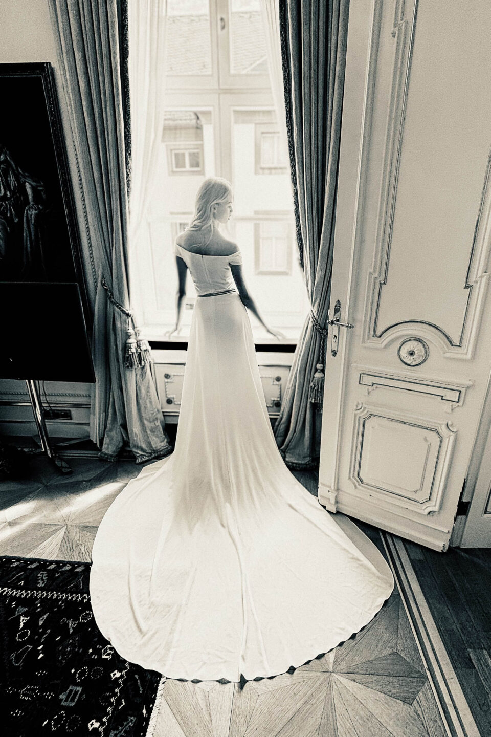 Schwarz-Weiß-Aufnahme Brautkleid Carlow, Rückenansicht mit langer Schleppe am Fenster