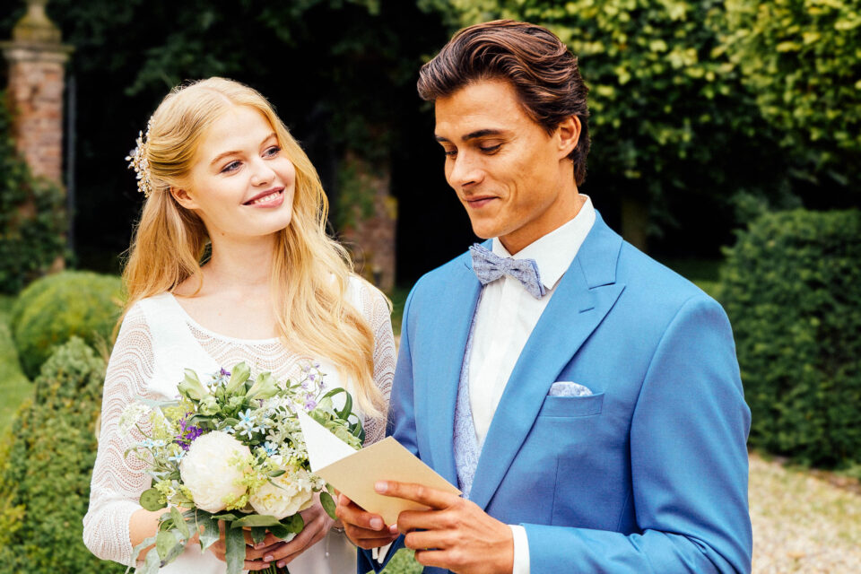 Brautpaar im Garten, Braut im Brautkleid Bobigny mit 3/4-Arm aus Wellenspitze, Bräutigam im blauen Anzug
